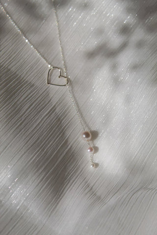Silver lariat necklace with heart-shaped pendant and three pearl drops over textured surface