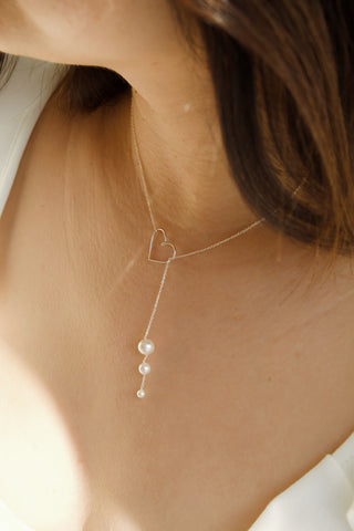 Silver lariat necklace with open heart pendant and three pearl drops on a person's neck.
