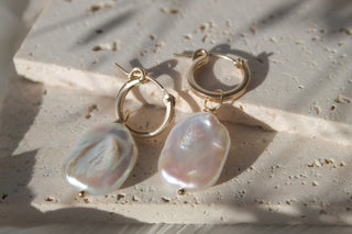 Close up of two small gold hoop earrings with baroque pearl drops on a textured stone surface