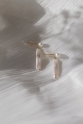 Pair of minimalist gold pearl stud with an elongated pearl drop earring on a white textured background. 